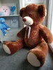 A large brown teddy bear is sitting on the floor in front of a white shelf with other stuffed animals and decorative items. Podgląd zdjęcia produktu