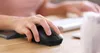 A person's hand is using a black computer mouse on a wooden desk. Voir les images du produit