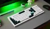 A white mechanical keyboard with black and teal keycaps is placed on a gray desk, with a green LED light bar in the background. Produktbild-Vorschau