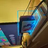 A clear acrylic case with blue LED lighting is shown holding Nintendo Switch game cards. The case is placed on a shelf next to a television screen displaying a menu. Produktbild-Vorschau