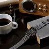 A watch, coffee cup, and a small wooden object are arranged on a dark surface, with a wooden speaker in the background. Produktbild-Vorschau