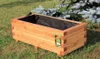 A wooden planter box sits on grass next to a pine tree. Produktbild-Vorschau