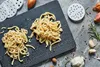 A top-down view of homemade noodles on a dark slate board, surrounded by ingredients like garlic, rosemary, and spices. Produktbild-Vorschau