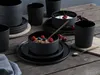 A collection of black stoneware dishes and cups arranged on a wooden table, with some dishes containing food. Produktbild-Vorschau