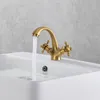 A brass faucet with two handles is mounted on a white sink, with water flowing from the spout into the sink. Náhled obrázku produktu