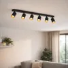 A black, six-light ceiling spotlight fixture is mounted on a white ceiling in a living room. The spotlights are angled downwards, illuminating the space below. A sofa and a shelf with plants are visible in the background. Náhľad obrázku produktu