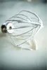 A whisk attachment with some white residue, likely meringue or whipped cream, is resting on a light-colored, scratched surface. Produktbild-Vorschau