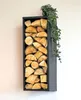 A black metal firewood rack filled with logs, mounted on a light-colored wall. A green plant drapes over the top right corner of the rack. Produktbild-Vorschau