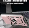 A rose gold smartphone case with a ring holder is shown at an angle, resting on a textured surface. The background is blurred, showing parts of a laptop. Produktbild-Vorschau