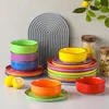 A collection of colorful ceramic bowls and plates are arranged on a white surface, with a decorative gray arch in the background. Produktbild-Vorschau
