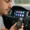 A man is holding a USB adapter in a car, with the car's infotainment screen visible in the background. Produktbild-Vorschau