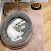 A white dog is resting in a round pet bed on a pink rug next to a food bowl. Produktbild-Vorschau