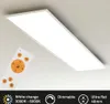 A white rectangular LED panel light is mounted on a light gray ceiling, with a white remote control in the foreground. The image highlights the product's features and specifications. Náhľad obrázku produktu