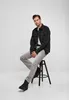 A man is sitting on a stool wearing a black jacket and gray jeans. Produktbild-Vorschau