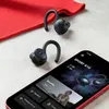 A pair of black wireless earbuds and a smartphone displaying the product's app are placed on a light gray surface next to a red cloth. Produktbild-Vorschau