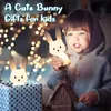 A child is holding a bunny-shaped night light, with another bunny night light in a gift box in the foreground. The background is blurred with bokeh lights. Produktbild-Vorschau