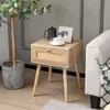 A wooden nightstand with a rattan drawer front is placed next to a sofa, with a plant in a pot to the left. Produktbild-Vorschau