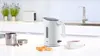 A white electric kettle is shown on a kitchen counter next to a cup of tea and some pastries. Produktbild-Vorschau
