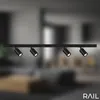 A black track light fixture with four adjustable spotlights is mounted on a black rail. The background is a blurred interior scene, likely a living room or dining area. Anteprima dell’immagine prodotto