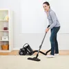 A woman is vacuuming a carpet with a canister vacuum cleaner in a brightly lit room. A bookshelf is visible on the left side of the image. Produktbild-Vorschau