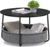 A round coffee table with a black top and a fabric basket underneath, containing a pillow, a blanket, and books. A potted plant sits on the table. Produktbild-Vorschau