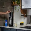 A person is using a SodaStream machine in a kitchen setting, with a bottle of Pepsi syrup and a glass of soda nearby. Produktbild-Vorschau
