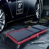 A red and black mechanic's creeper is positioned in front of a dark blue car in a garage setting. Náhľad obrázku produktu