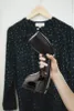 A hand holding a black handheld garment steamer in front of a dark patterned blouse on a hanger. Voir les images du produit