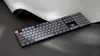 A dark gray keyboard with an orange escape key is placed on a dark surface with a white paper-like object curving behind it. Produktbild-Vorschau