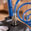 A close-up view of an air compressor's connection point, showing a blue hose with a spring guard attached to a silver quick-connect fitting, which is plugged into the black compressor unit. A pressure gauge is partially visible on the left. Anteprima dell’immagine prodotto