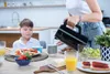 A person is pouring water from a black electric kettle into a mug at a breakfast table with a child. Anteprima dell’immagine prodotto