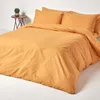 A bed with orange bedding and two pillows, with a small plant on a side table in the background. Produktbild-Vorschau