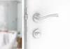 A close-up view of a silver door handle and lock mechanism on a white door, with a blurred bathroom scene visible through the open doorway. Produktbild-Vorschau