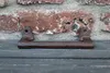 A rusty metal handle sits on a dark wooden surface in front of a brick wall. Produktbild-Vorschau