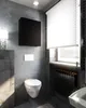 A modern bathroom with concrete walls, a wall-mounted toilet, a black cabinet, and a radiator under a window with blinds. Produktbild-Vorschau