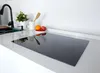 A modern kitchen countertop with a black induction cooktop in the foreground, a sink and faucet to the left, and a small fruit bowl and plant in the background. Produktbild-Vorschau