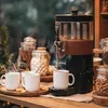 A hot chocolate dispenser is set up on a wooden counter, with mugs, cookies, and jars of toppings nearby, suggesting a cozy winter or holiday setting. Podgląd zdjęcia produktu