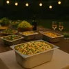 An outdoor dinner party spread with various food dishes served in metal trays, with string lights in the background. Produktbild-Vorschau