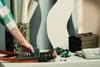 A hand is using a green stapler on a wooden object, surrounded by rolls of paper, fabric, and a toolbox. Podgląd zdjęcia produktu