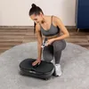 A woman is kneeling on a rug next to a vibration plate, holding a remote control. Náhľad obrázku produktu