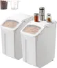Two white food storage containers with clear lids, filled with pasta, are shown. Small spice jars and a pepper grinder are on top of one container. In the background, two clear measuring cups are visible, one filled with small brown items. Náhled obrázku produktu