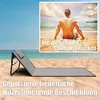 A beach scene with a lounge chair and a man sitting on the sand, looking out at the ocean. Produktbild-Vorschau