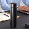 A black smart LED thermobecher is placed on a desk next to a notebook and pen, with a person typing on a laptop in the background. Náhľad obrázku produktu