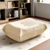 A beige floor cushion is placed on a rug in a room with bookshelves in the background. Náhled obrázku produktu