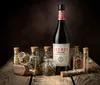A bottle of Lustau Vermut Red is displayed prominently on a rustic wooden surface, surrounded by various small glass bottles filled with spices and herbs. The background is a dark, textured brown. Produktbild-Vorschau