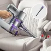 A hand is using a silver and purple vacuum cleaner to clean a white car seat. A close-up of the vacuum attachment is shown in a circle. Produktbild-Vorschau
