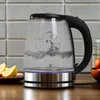 A glass electric kettle with boiling water on a kitchen counter, next to lemons and a bowl of fruit. Produktbild-Vorschau
