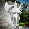 A white outdoor wall lamp is mounted on a stone wall, with a garden and pool in the background. Náhľad obrázku produktu