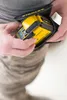 A person is putting a yellow and black Stanley laser level into a black belt pouch. Produktbild-Vorschau