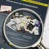 A magnifying glass is placed over a collection of documents and a smartphone, suggesting a crime investigation or puzzle-solving scenario. Produktbild-Vorschau